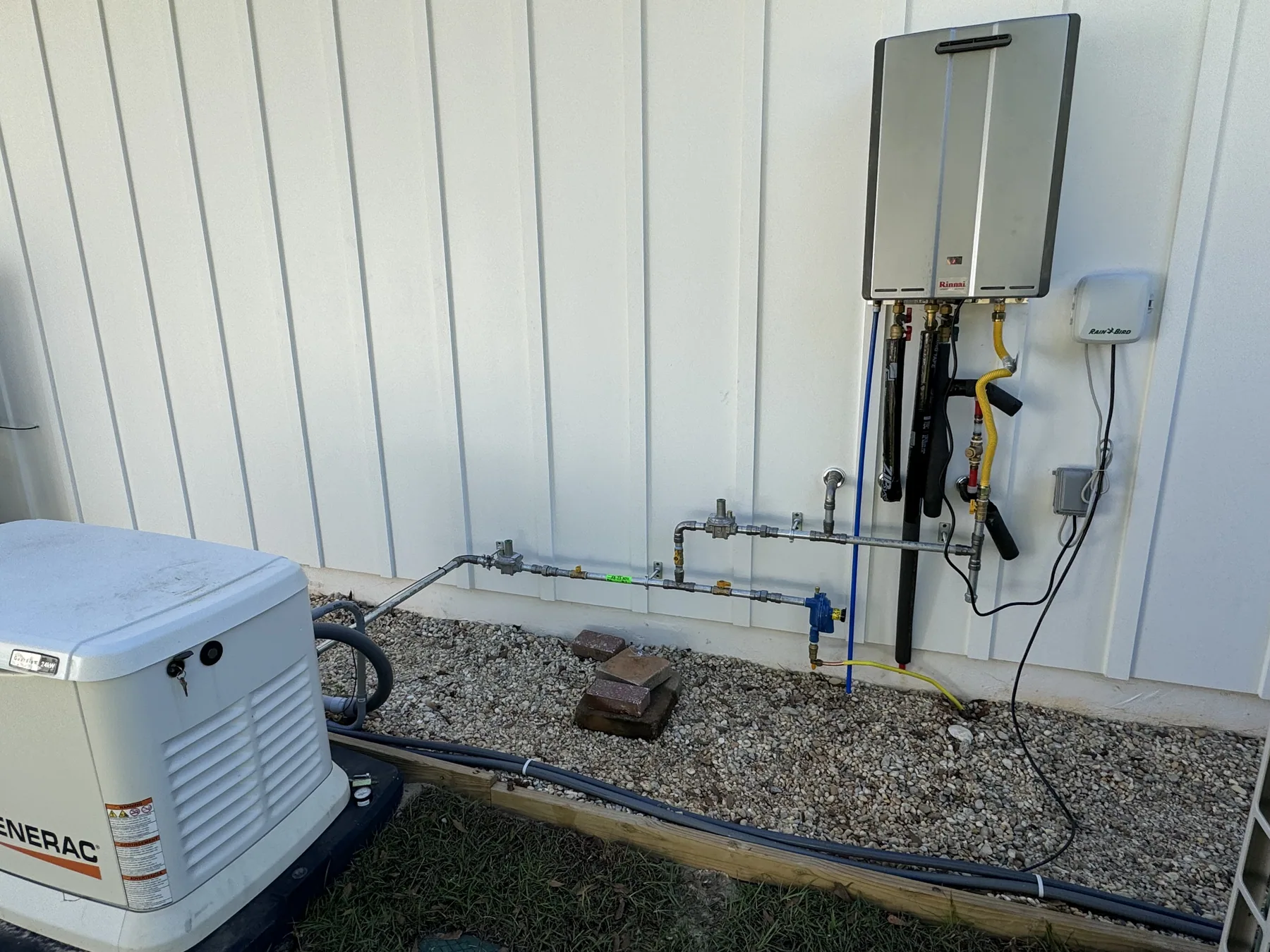 A Rinnai tankless water heater mounted on the white board-and-batten wall of a house, its gas and water lines running down to a gravel bed beside a standby generator