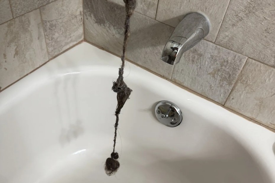 A tub drain stopper lifted out of a bathtub with a long strand of hair hanging from it