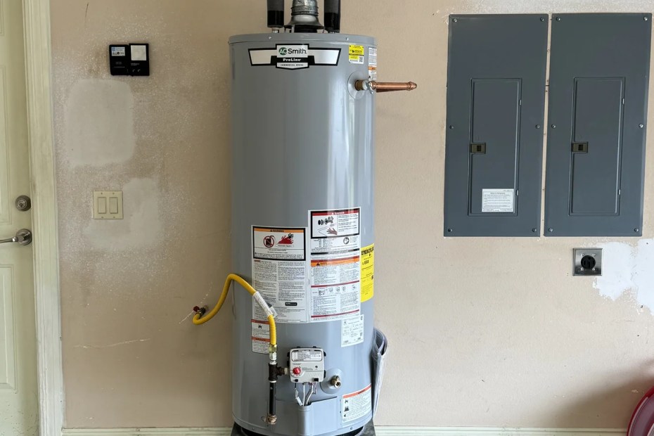 A gas tank water heater on a metal stand in a garage, vent pipe and expansion tank above it, electrical panels beside it